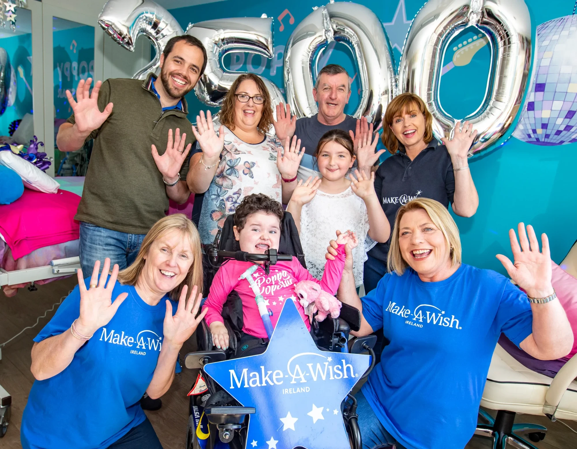 A young wish child in a wheelchair celebrating with family and Make-A-Wish Ireland volunteers, all waving under silver 2500 balloons